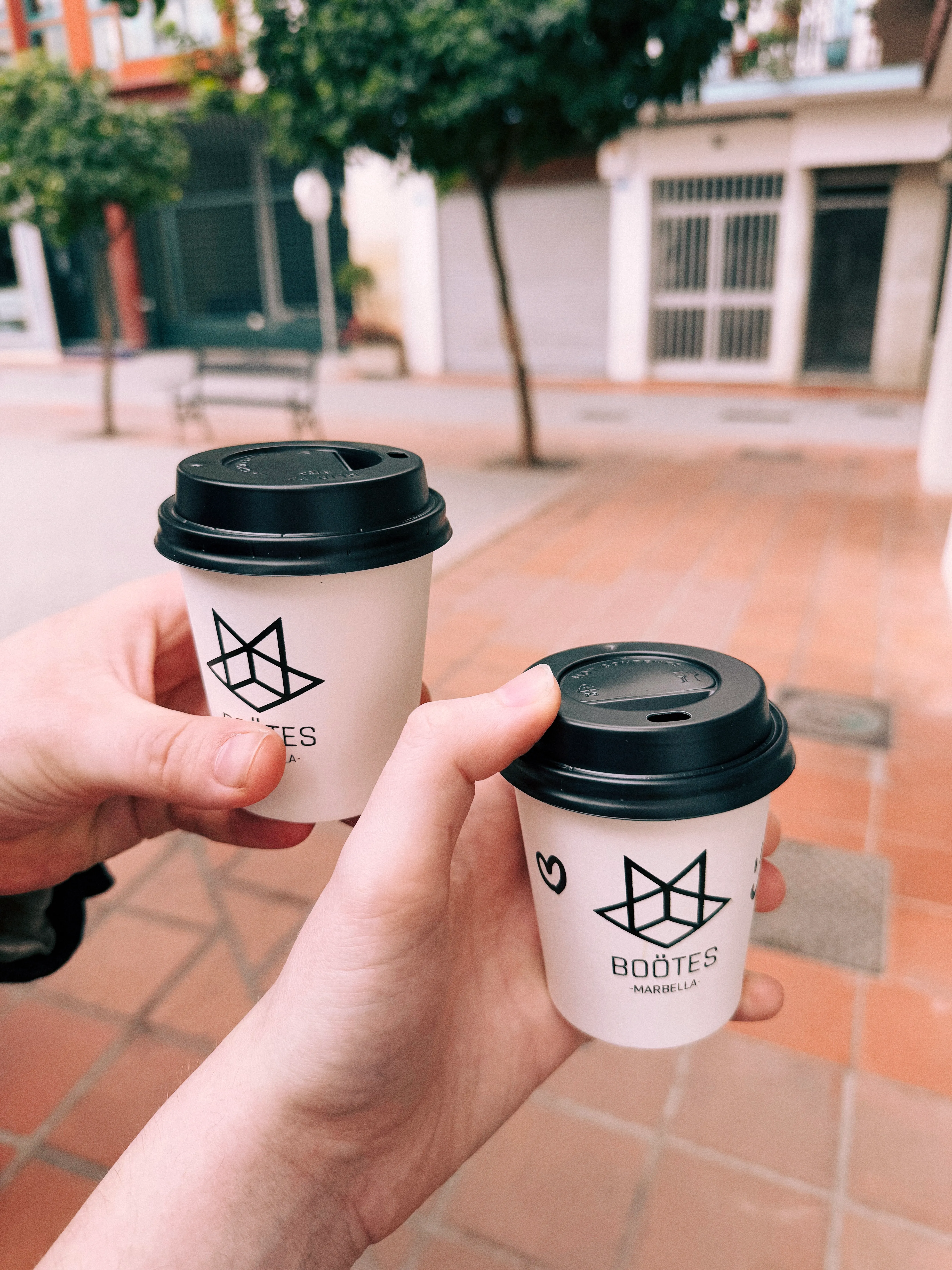 Two hands, each holding a coffee cup that says "Böotes Marbella". The cup on the right has a heart drawn on it.